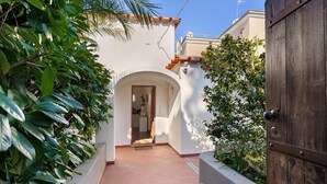 A narrow pathway leading to a doorway, surrounded by lush greenery and a white building with a tiled roof.