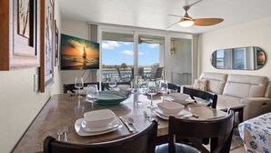 Un coin repas avec une table dressée pour quatre personnes, un ventilateur de plafond et un tableau représentant un coucher de soleil sur l’eau.
