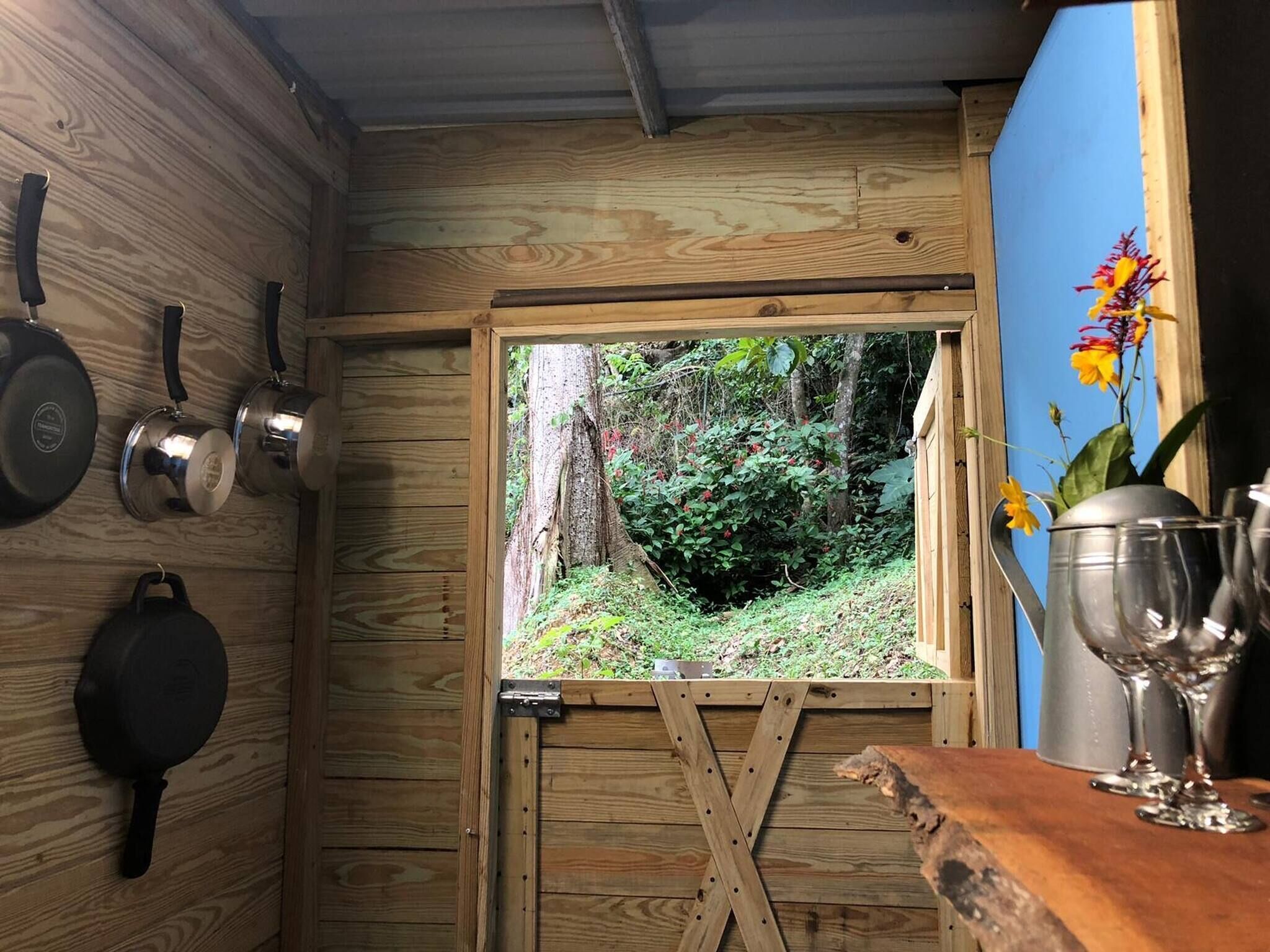 A rustic wooden interior with a window view of a forest, a wooden counter with kitchen utensils, and a metal pot on the right.