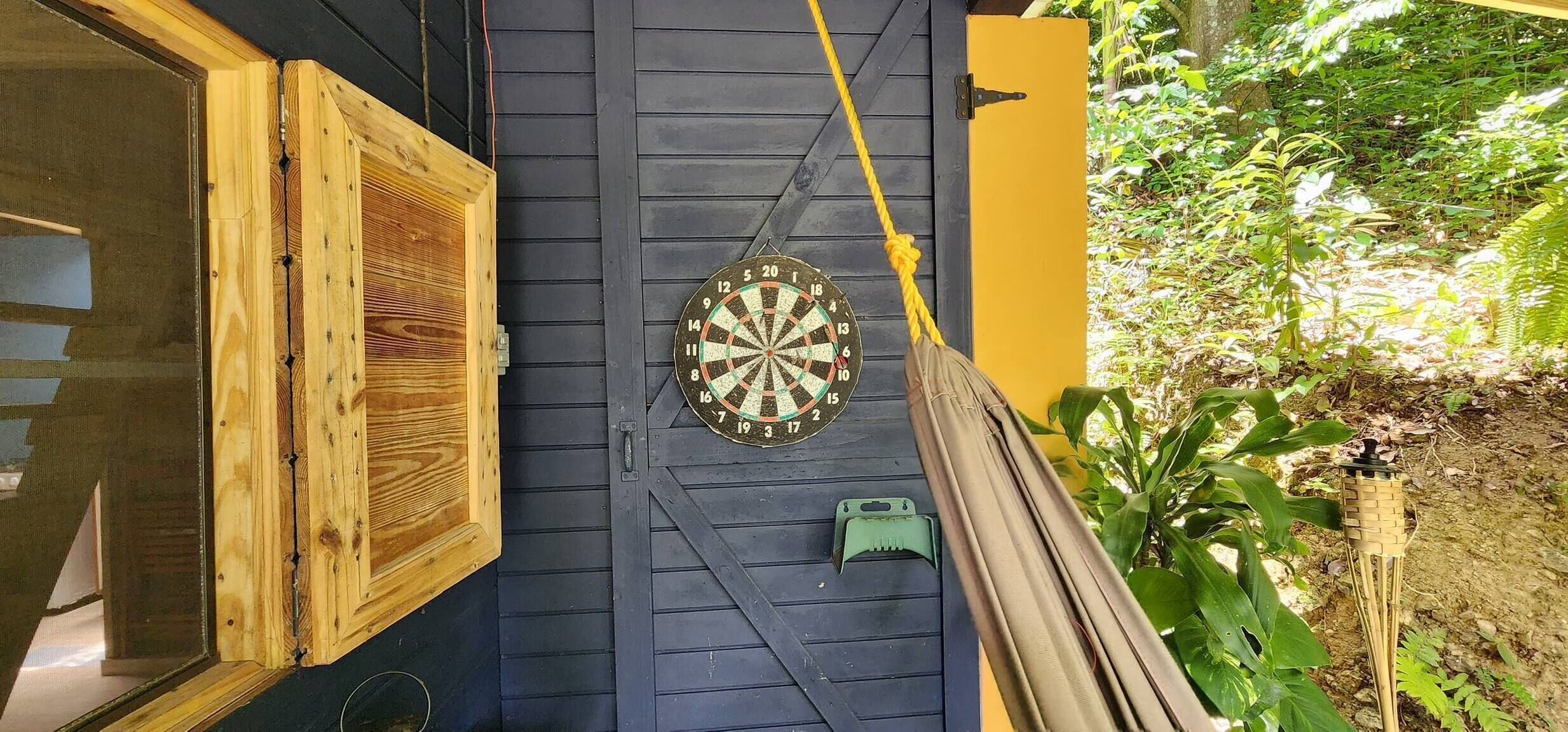 Un bersaglio di freccette fissato a una parete di legno scuro, una finestra di legno con una persiana socchiusa e un'amaca legata al muro.