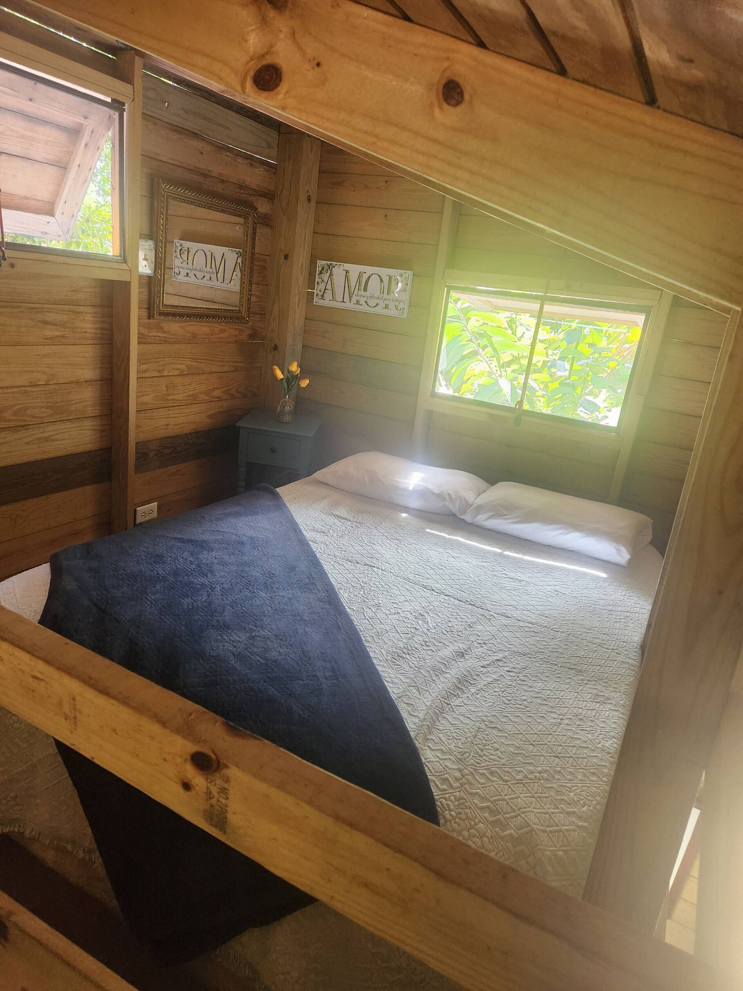A wooden cabin bedroom with a bed, a nightstand with a vase of flowers, and a window with a view of greenery.