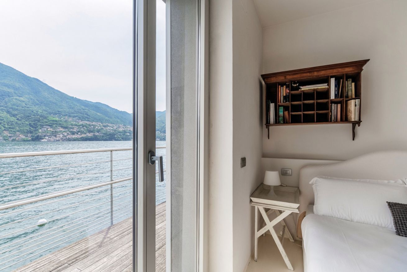 Een slaapkamer met uitzicht op een meer en bergen, een houten boekenkast, een tafeltje met een lamp, en een wit bed.