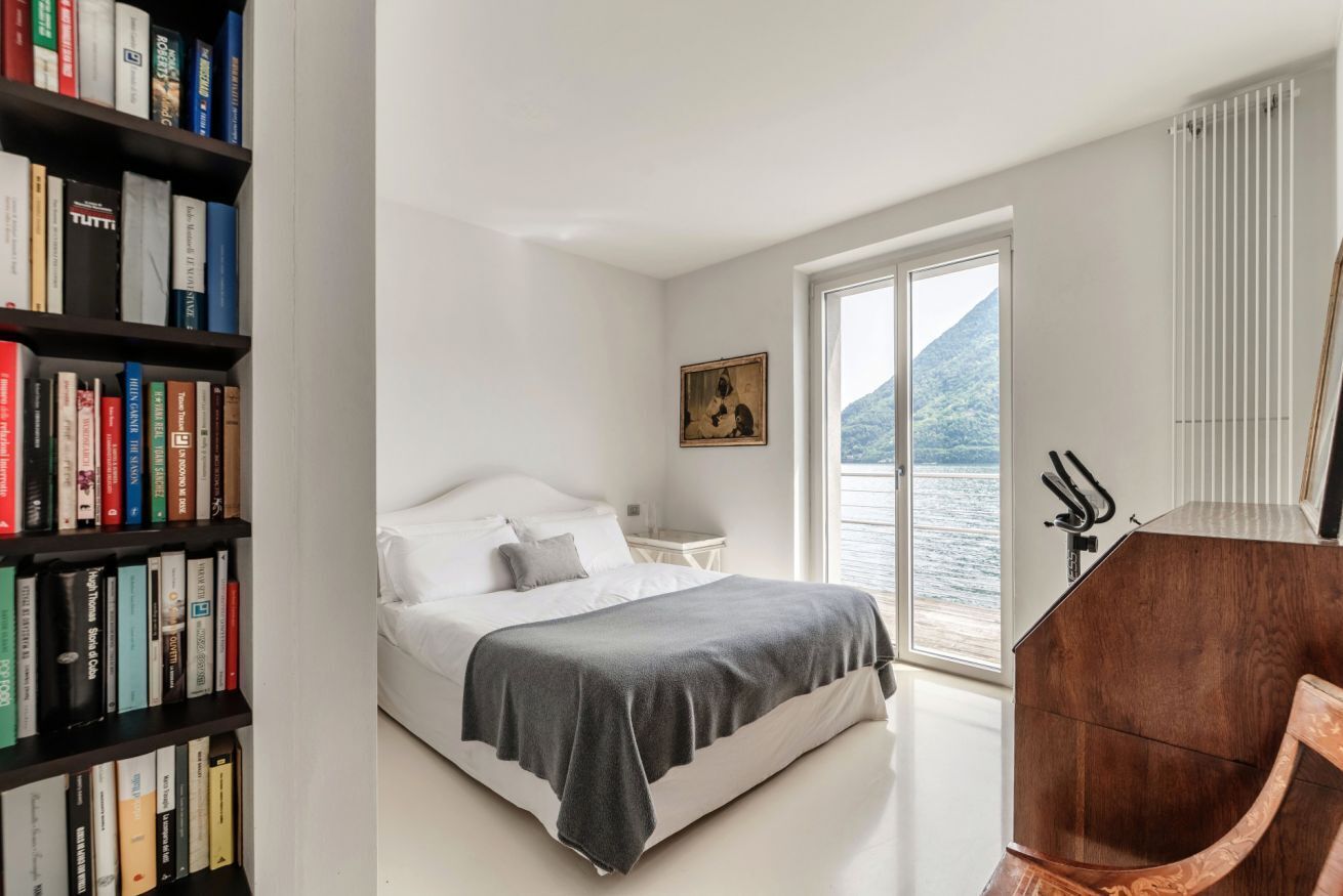 A bedroom with a bed, a bookshelf, a painting, a window with a view of water, and a wooden chair.
