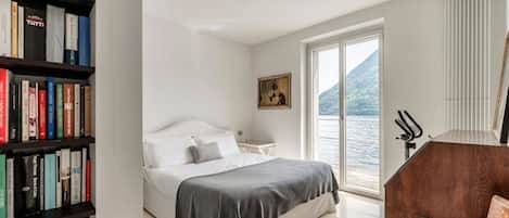 A bedroom with a bed, a bookshelf, a painting, a window with a view of water, and a wooden chair.