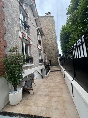 A narrow outdoor corridor with a tiled floor, a potted plant, and a chair.