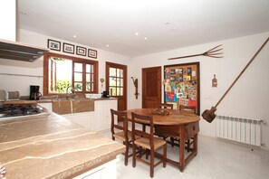 Een keuken met een kookplaat, een eettafel met stoelen, houten kastjes en een prikbord aan de muur.