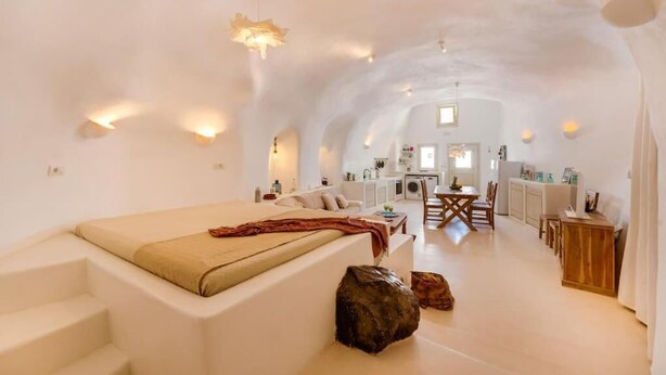 A spacious, white-walled room with a bed, a dining area, and a kitchen. - Cave at White Rock  Private  jacuzzi  (Santorini)