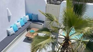 A sunlit outdoor seating area with a palm tree, a small table, and a white wall. - Cave at White Rock  Private  jacuzzi  (Santorini)