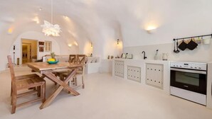 A modern kitchen with white cabinetry, a wooden dining table, and a hanging light fixture. - Cave at White Rock  Private  jacuzzi  (Santorini)