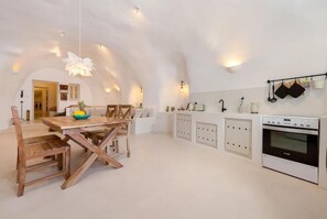 A modern kitchen with white cabinetry, a wooden dining table, and a hanging light fixture. - Cave at White Rock  Private  jacuzzi  (Santorini)