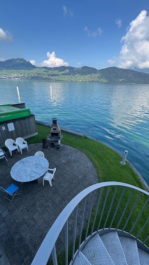 Een terras met buitenmeubilair, een barbecue en uitzicht op een meer met bergen op de achtergrond.