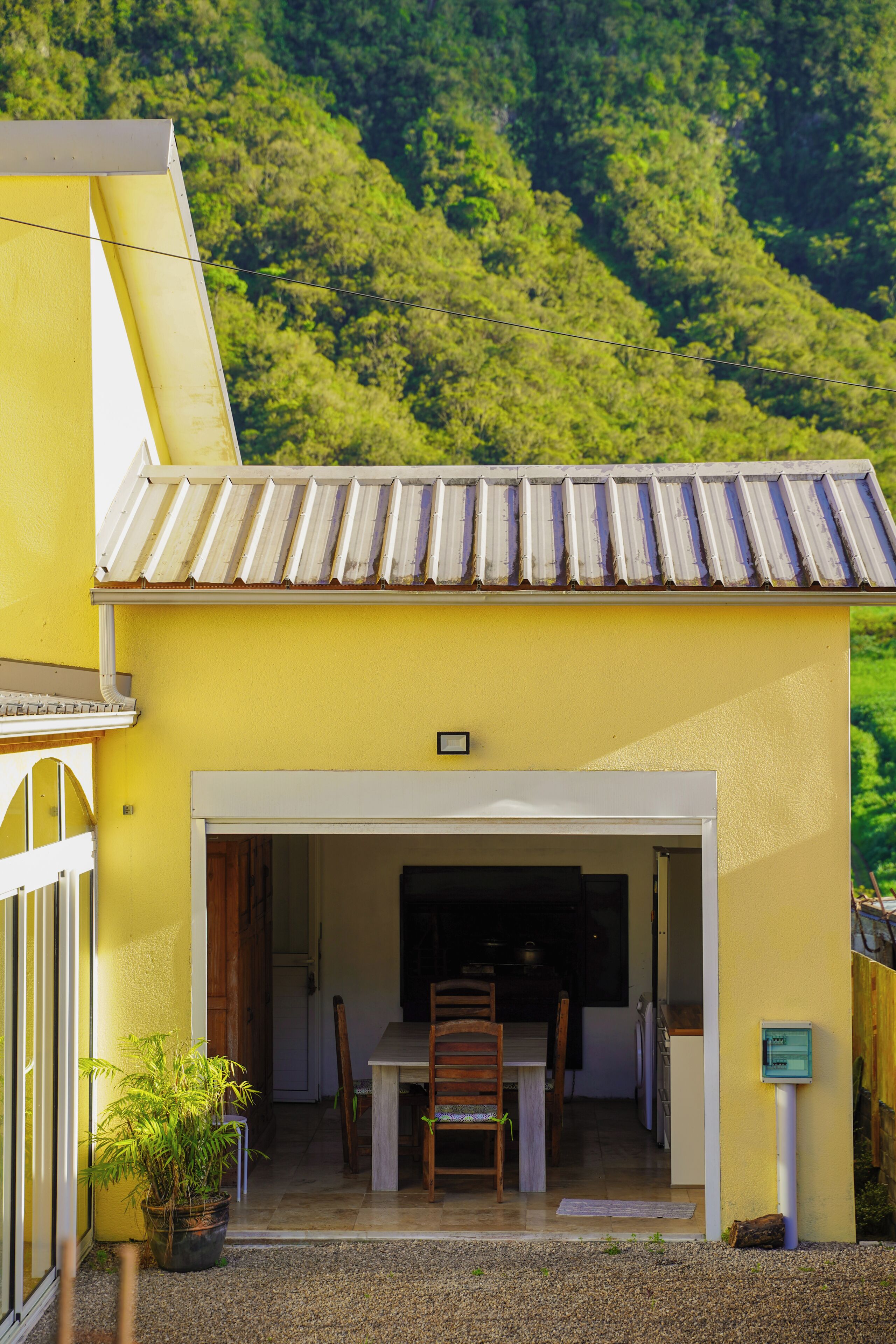 A yellow building with a tiled roof, a dining area with chairs and a table, and a potted plant outside.