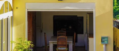 A yellow building with a tiled roof, a dining area with chairs and a table, and a potted plant outside.