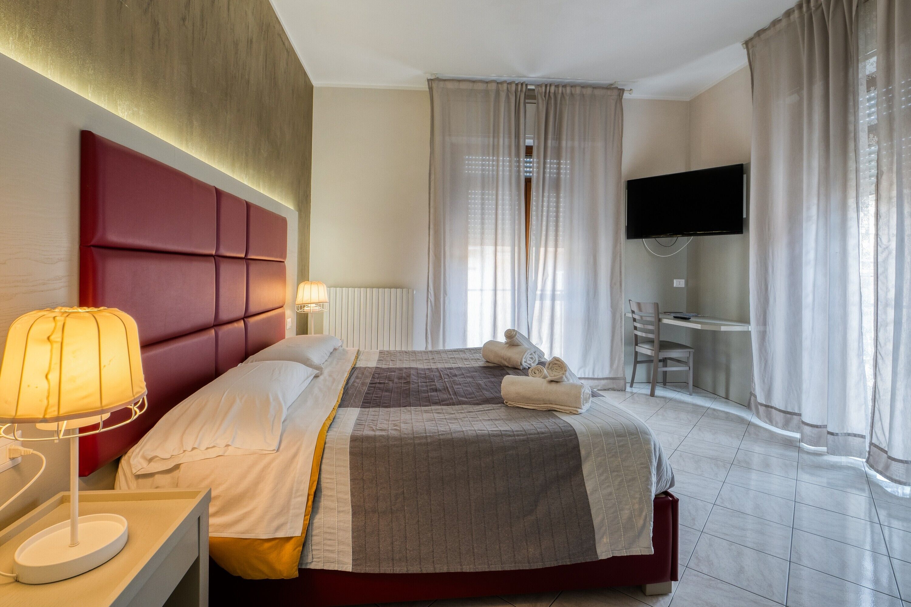 A hotel room with a bed, bedside lamp, curtains, a TV, and a desk.