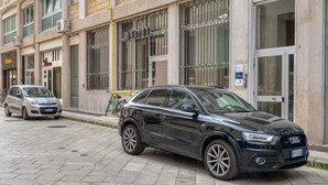 Um carro preto estacionado na rua pavimentada em frente a um edifício com janelas protegidas por grades e uma placa com a inscrição 'IBNL'.