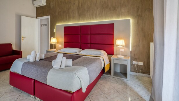 Quarto de hotel com cama vermelha, duas mesas de cabeceira com luminárias, um sofá vermelho e uma parede de madeira.
