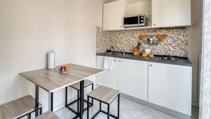 Uma cozinha compacta com micro-ondas, pia e painel de azulejos. Há duas mesas de madeira com bancos, e uma pequena prateleira com utensílios de cozinha.