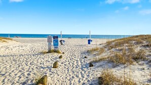 Una playa arenosa con cielo azul despejado, algunas personas a lo lejos y un cartel informativo.