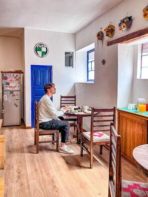 A person seated at a wooden dining table with chairs, a refrigerator with magnets, a blue door, and a window with a view. - Flying Dog Hostel Cusco (Cusco)