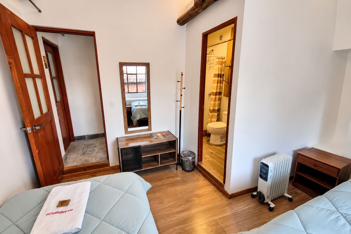 Quarto compacto com cama, uma secretária pequena, radiador e banheiro visível por uma porta aberta.
