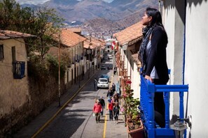 Uma rua estreita ladeada por edifícios, uma pessoa em uma varanda, e uma paisagem montanhosa ao fundo.
