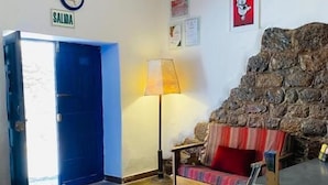 A room with a wooden floor, a blue door labeled 'SALIDA', a lamp, a sofa with a red cushion, and a stone wall. - Flying Dog Hostel Cusco (Cusco)