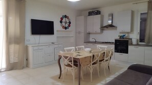 A modern kitchen with white cabinets, a dining table with chairs, a flat-screen TV, and a decorative wall clock.