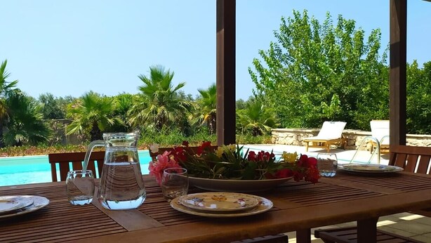 Une table en bois, sur laquelle se trouvent une carafe, des assiettes et des verres, donnant sur une piscine bordée de palmiers et sur un mur de pierre en arrière-plan.