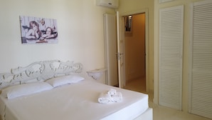 Une chambre avec un lit blanc, un climatiseur fixé au mur, une porte avec une fenêtre et un radiateur blanc.