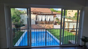 Une pièce dotée d’une grande porte-fenêtre coulissante donnant sur un jardin avec une piscine, une terrasse en briques et une vue sur les maisons voisines.