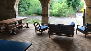 Une terrasse extérieure dotée de mobilier en bois, d’arches en pierre et offrant une vue sur la verdure.