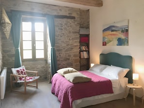 Un dormitorio con pared de piedra, cama con cabecera verde azulada, silla con cojín de lunares, estantería y un cuadro en la pared.