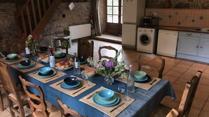 On aperçoit une salle à manger avec une longue table dressée pour six personnes, entourée de chaises en bois. Au fond, on distingue une cuisine avec un mur en pierre et un mur arrière recouvert de carreaux.