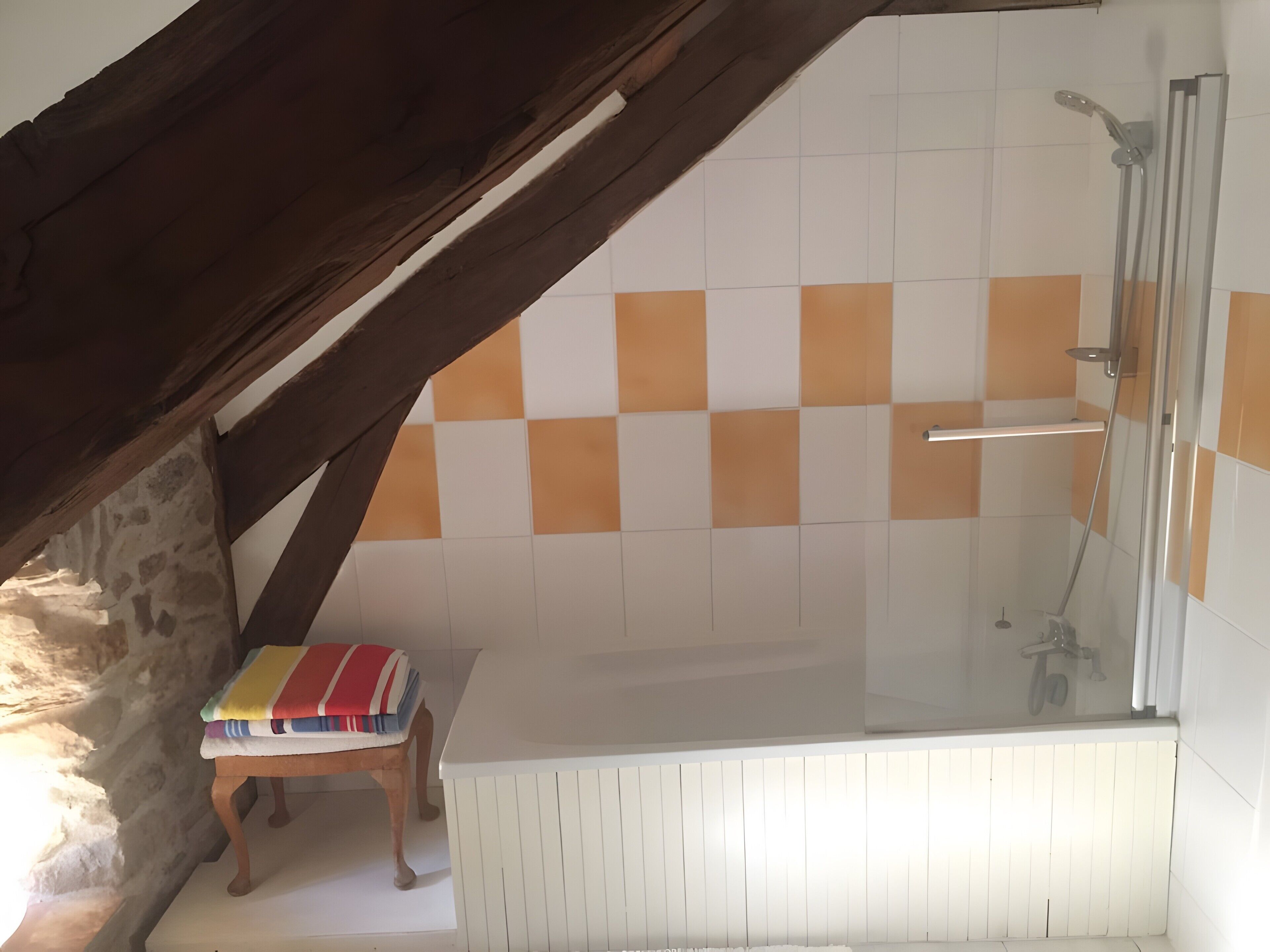 A bathroom with a shower, a tiled wall, and a wooden stool with a striped towel.