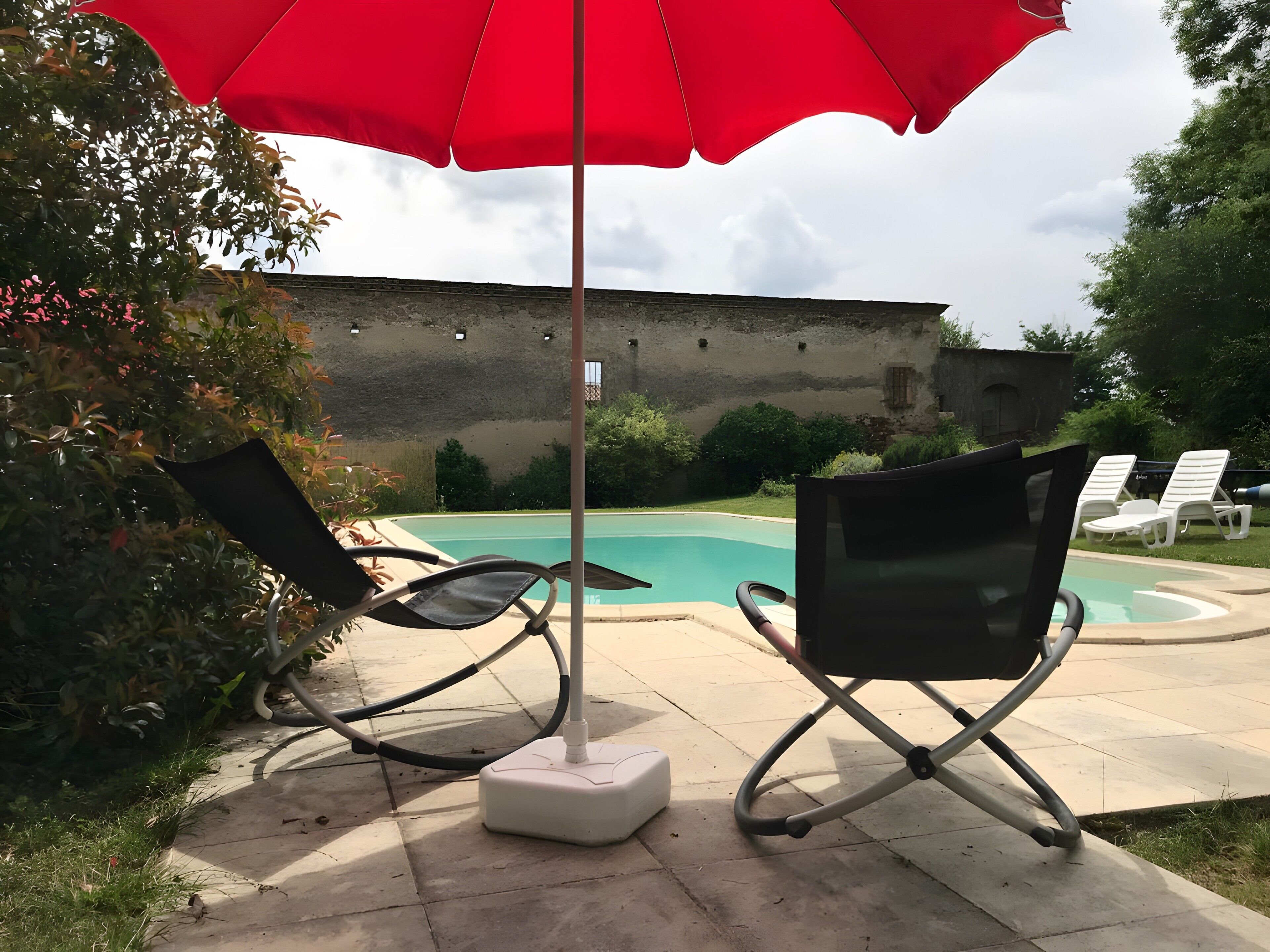 A poolside area with two lounge chairs, a red umbrella, and a stone wall in the background.