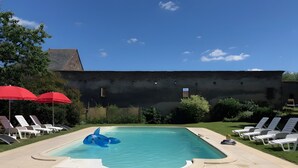 Une piscine entourée de verdure et d’un bâtiment, avec des chaises longues et des parasols rouges.
