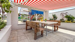Un patio al aire libre con un toldo a rayas rojas y blancas, un banco de madera, una mesa y plantas en macetas.