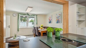 Una cocina moderna con una isla de vidrio, vigas de madera, un comedor con mesa y sillas, y plantas en macetas.