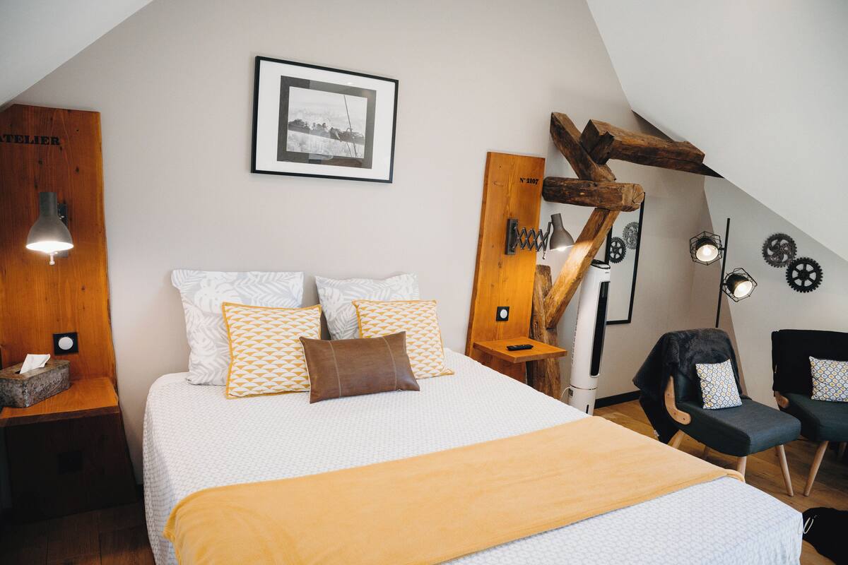 Une chambre à coucher avec un grand lit, une tête de lit en bois, un tableau encadré au mur et une chaise avec un coussin.