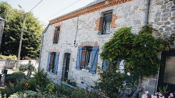 Un bâtiment en pierre avec des volets bleus, entouré d’une végétation luxuriante et de plantes en fleurs.