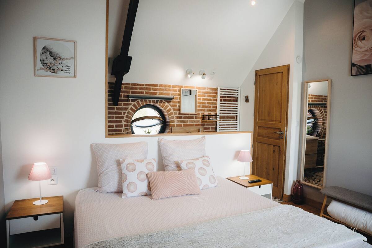 Une chambre avec un mur en briques, une porte en bois, un lit agrémenté d’oreillers à motifs, des tables de chevet avec des lampes et un miroir sur le mur.