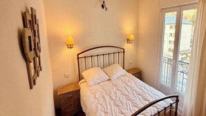 Ein Schlafzimmer mit hölzernem Bettgestell, zwei Nachttischen, einer Wandlampe und einem Fenster mit Blick auf Gebäude.