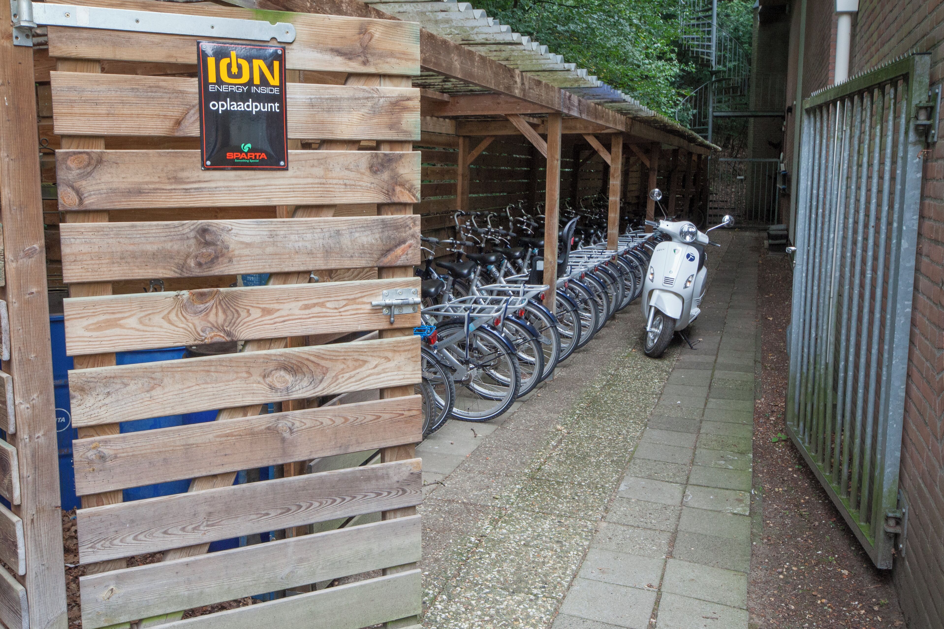 Una estructura de madera con un letrero que dice "ION ENERGY INSIDE opladpunt" y una fila de bicicletas estacionadas bajo un área techada.