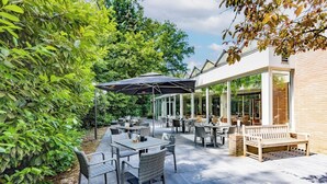 An outdoor patio area with tables and chairs, surrounded by greenery and a building with large windows. - Peaceful Getaway — Free Parking, Indoor Pool, and Everything You Need to Unwind (Lochem)