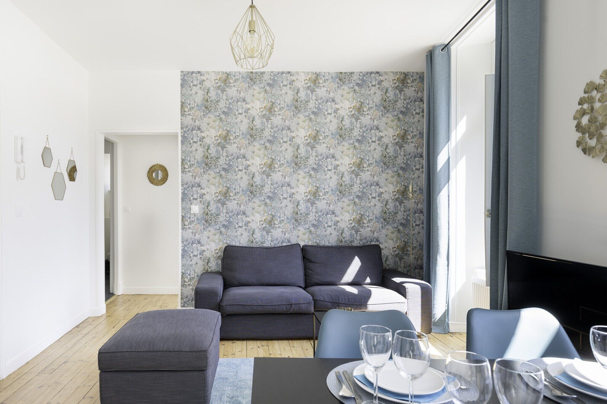Ein modernes Wohnzimmer mit einem grauen Sofa, einem Esszimmertisch gedeckt mit Tellern und Gläsern sowie Tapete mit Muster.