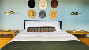 A bedroom with a bed, a plaid pillow, and a wall with various hats. There are two bedside tables with lamps. - Historic Store & Basecamp for Fort Davis, Marfa, Alpine and all things Far West (Fort Davis)