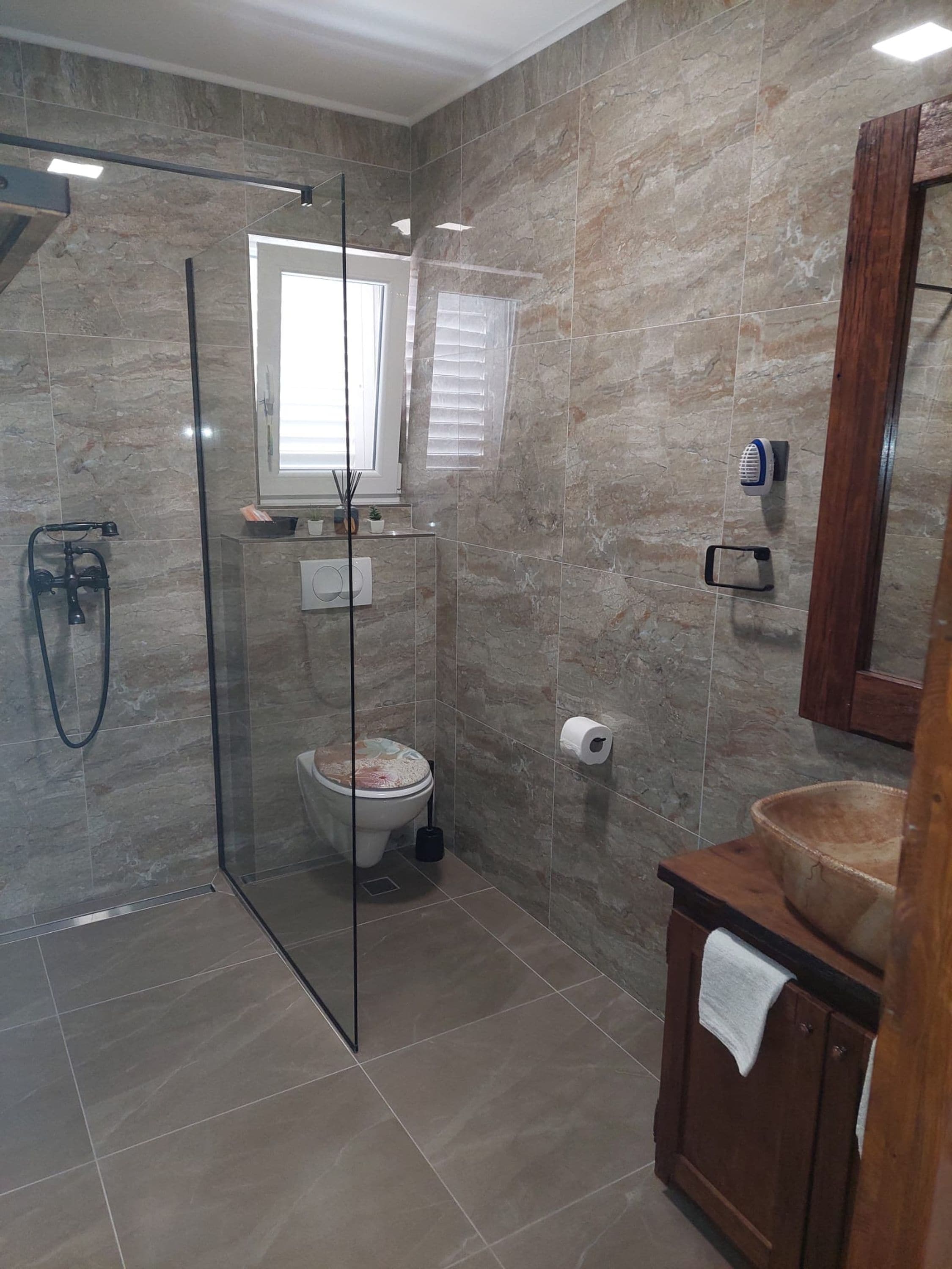 A modern bathroom with a glass-enclosed shower, a toilet with a decorative cover, a wooden vanity with a bowl sink, and a mirror above the vanity.