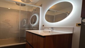 Ein modernes Badezimmer mit einer Duschkabine aus Glas, einem weißen Waschbecken und einem runden Spiegel mit beleuchtetem Rahmen.