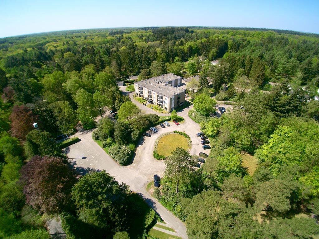 An aerial view of a hotel surrounded by dense forest, with a central parking area and a building with multiple floors.