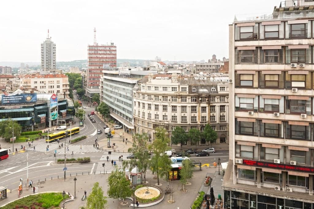 Paisagem urbana com uma mistura de edifícios modernos e históricos, uma rotatória central e um ponto de ônibus.